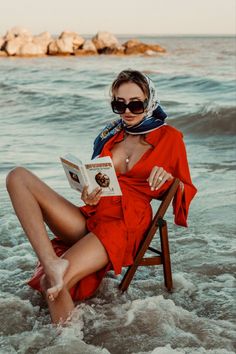 Woman in a red dress and head scarf sitting on a chair in the ocean reading a book
