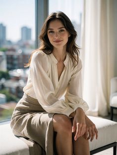 Woman with long hair in a blouse and midi skirt sitting indoors with a cityscape view
