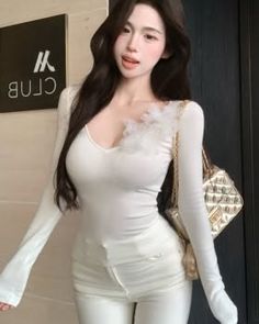 Woman with long dark hair wearing a white long-sleeve top and white pants indoors