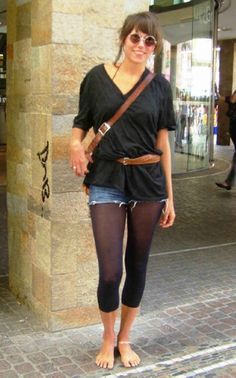 Woman in round sunglasses, denim shorts, and tights, standing barefoot on a street