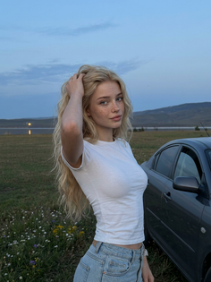 Blonde woman with freckles in a white t-shirt and jeans outdoors with a car