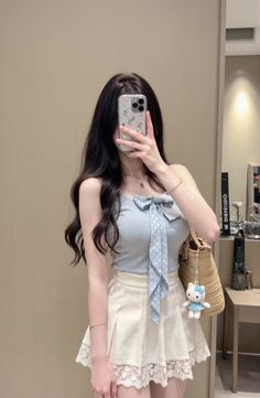 Woman with long dark hair taking a mirror selfie in a light blue top and white skirt