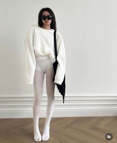 Woman in a white hoodie, white tights, and black sunglasses standing indoors