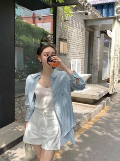 Woman in a striped shirt and white shorts drinking from a cup on a sunny street