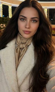 Woman with long dark hair and green eyes wearing a white coat and beige scarf at night
