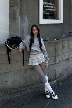 Woman in a grey sweatshirt and ruffled skirt leaning against a wall outdoors