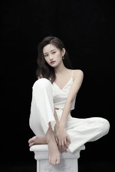 Woman in a white top and pants sitting on a white pedestal against a dark background