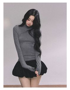 Woman with long black hair wearing a grey turtleneck and black skirt indoors