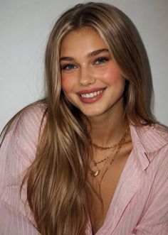 Smiling woman with long blonde hair and layered necklaces wearing a pink shirt