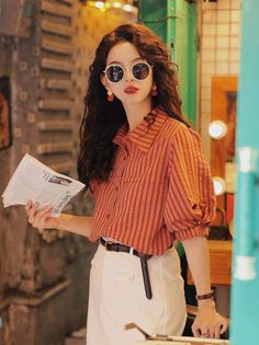 Woman with long hair and sunglasses wearing a striped shirt and white pants indoors