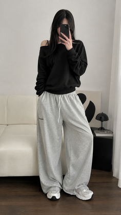 Woman in a black off-shoulder top and grey sweatpants taking a mirror selfie