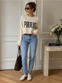 Woman in a Paris sweater and light wash jeans holding a brown bag indoors