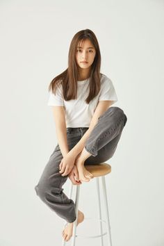 Woman with long hair in a white t-shirt and gray jeans sitting barefoot on a stool