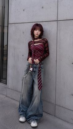 Woman with shaggy hair wearing a plaid top and baggy jeans against a concrete wall