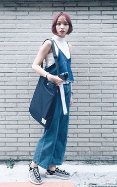 Woman with pink short hair wearing denim overalls and a white turtleneck against a brick wall