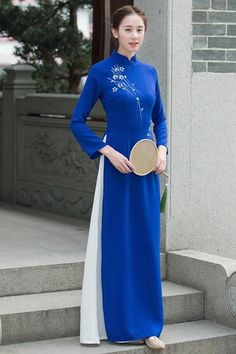 Woman in a blue ao dai with white embroidery holding a fan on stone steps