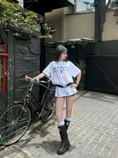 Woman in a white t-shirt, black shorts, and beanie posing with a bicycle outdoors