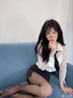 Woman with long black hair and glasses in a white shirt and mini skirt sitting on a couch