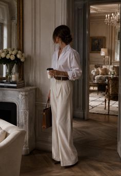 Woman in a white shirt and wide leg pants holding coffee in a classic interior