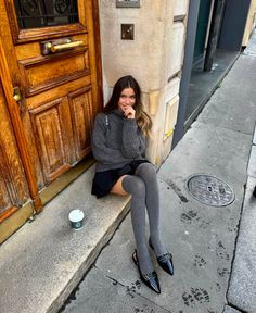 Woman in a gray sweater and black skirt sitting on a street curb