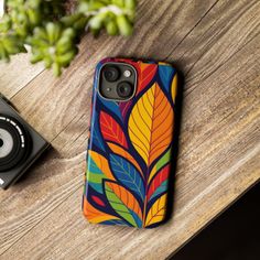 A phone case with a colorful leaf pattern on a wooden table next to a camera