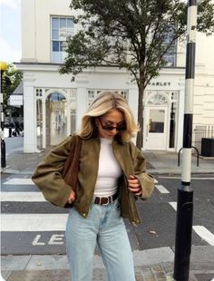 Woman with blonde hair and sunglasses wearing a green jacket and blue jeans on a street