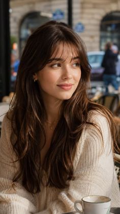 Woman with long brown hair and bangs wearing a cream sweater at an outdoor cafe