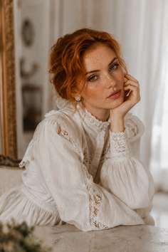 Woman with red hair and freckles wearing a white vintage-style dress with lace details