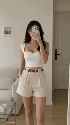 Woman in a white crop top and high-waisted shorts taking a mirror selfie indoors