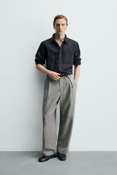 Man in a black shirt and grey pants posing against a white wall