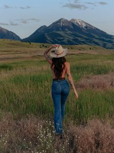 Woman in a cowboy hat and blue jeans walking through a field with mountains in the background