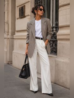Woman in a plaid blazer and white pants holding a black handbag outdoors