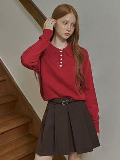 Woman with red hair and freckles wearing a red sweater and pleated skirt indoors
