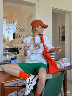 Woman in a white t-shirt and green shorts sitting on a desk indoors