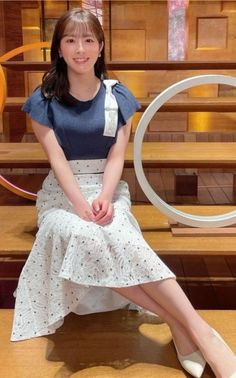 Woman with long hair and bangs wearing a blue top and white skirt, sitting indoors