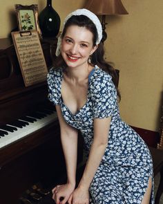 Woman in a blue floral dress and white headband posing by a piano