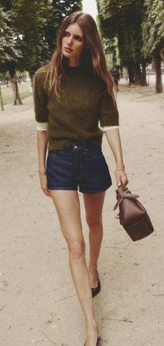 Woman with long hair wearing a green sweater and denim shorts walking outdoors