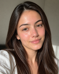 Woman with long brown hair in a white shirt, smiling in soft indoor lighting