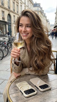 Woman in a trench coat smiling at an outdoor cafe with a glass of white wine