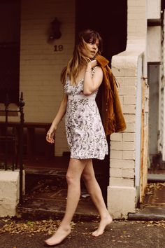 Woman in a short floral dress walking barefoot outdoors with a jacket over her shoulder