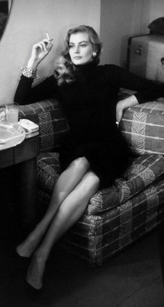 Woman in a black turtleneck dress sitting on a patterned couch, smoking a cigarette