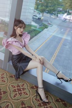 Woman in a pink top and black skirt sitting by a window overlooking a cityscape