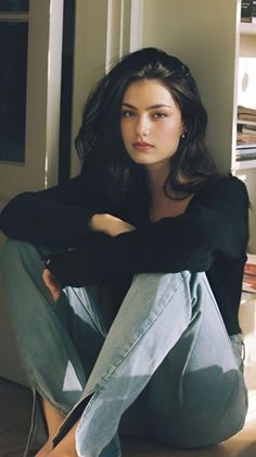 Woman with long dark hair wearing a black top and light wash jeans, sitting indoors