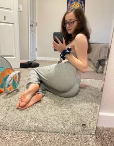 Woman in glasses and sweatpants taking a mirror selfie on a carpeted floor