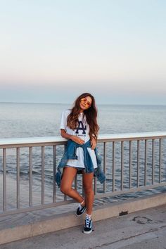 Woman in a white t-shirt and denim jacket leaning on a railing by the ocean