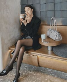 Woman in a black blazer and tights applying lipstick while sitting indoors