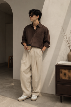 Man in a brown shirt and wide leg pants holding a coffee cup indoors