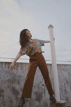 Woman in a patterned shirt and brown pants posing outdoors against a concrete wall
