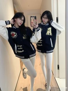 Two women in varsity jackets and white leggings taking a mirror selfie indoors