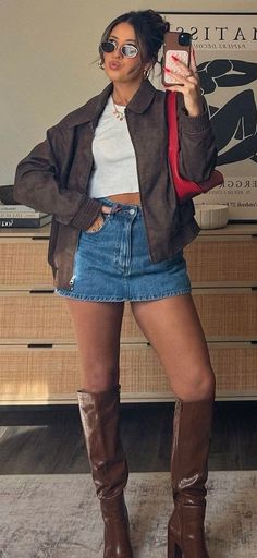 Woman in a denim skirt, brown jacket, and knee-high boots posing indoors